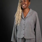 portrait image, merchant and mills Winnie pyjama pattern,woman standing, angled to right side, looking up, smiling. modelling long sleeve shirt, shorts, fabric is a light and dark grey stripe, background is black.