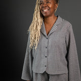 portrait image, merchant and mills Winnie pyjama pattern,woman standing, angled to right side, looking up, smiling. modelling long sleeve shirt, shorts, fabric is a light and dark grey stripe, background is black.