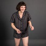 portrait image, merchant and mills Winnie pyjama pattern, woman stood, one foot unfront of the other, smiling, looking off camera, modelling short sleeve shirt, shorts, fabric is a navy and brown wide stripe, background is black.