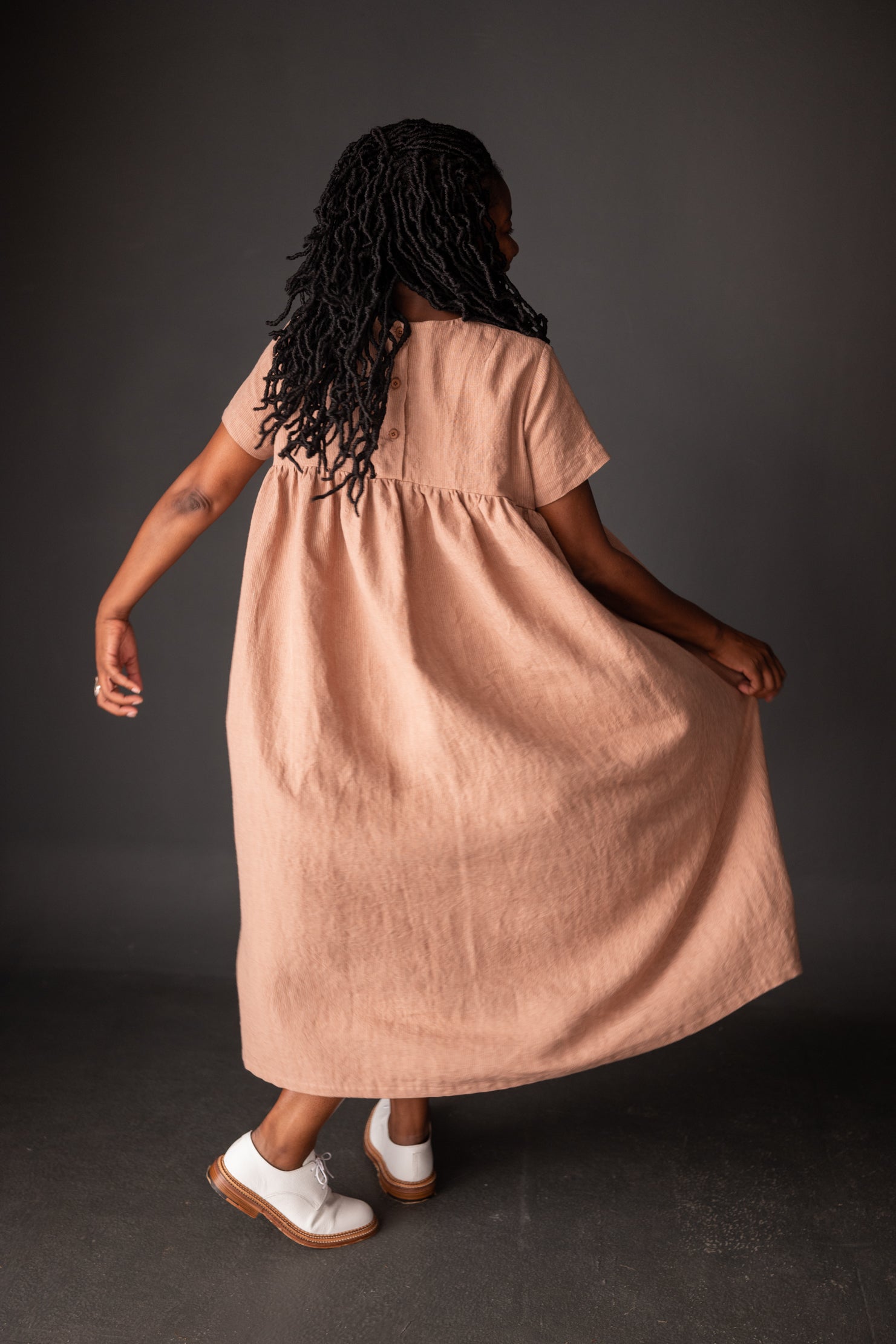 portrait image, merchant and mills Florence pattern, woman standing, facing back, looking ahead, right arm holding dressmodelling shin length dress, paired with white brogues, waist is gathered, bodice resembles a t-shirt styles. sleeves are short. fabric is peach background is black