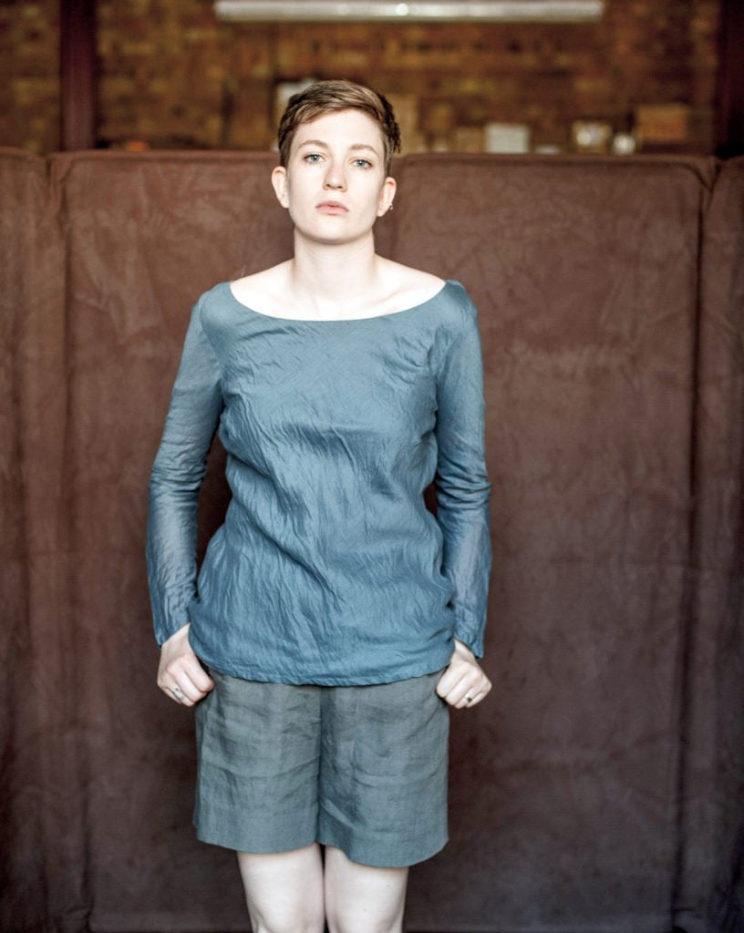 portrait image, woman stood face on, no emotion, thumbs in long grey shorts pocket, modelling teal long sleeve top.