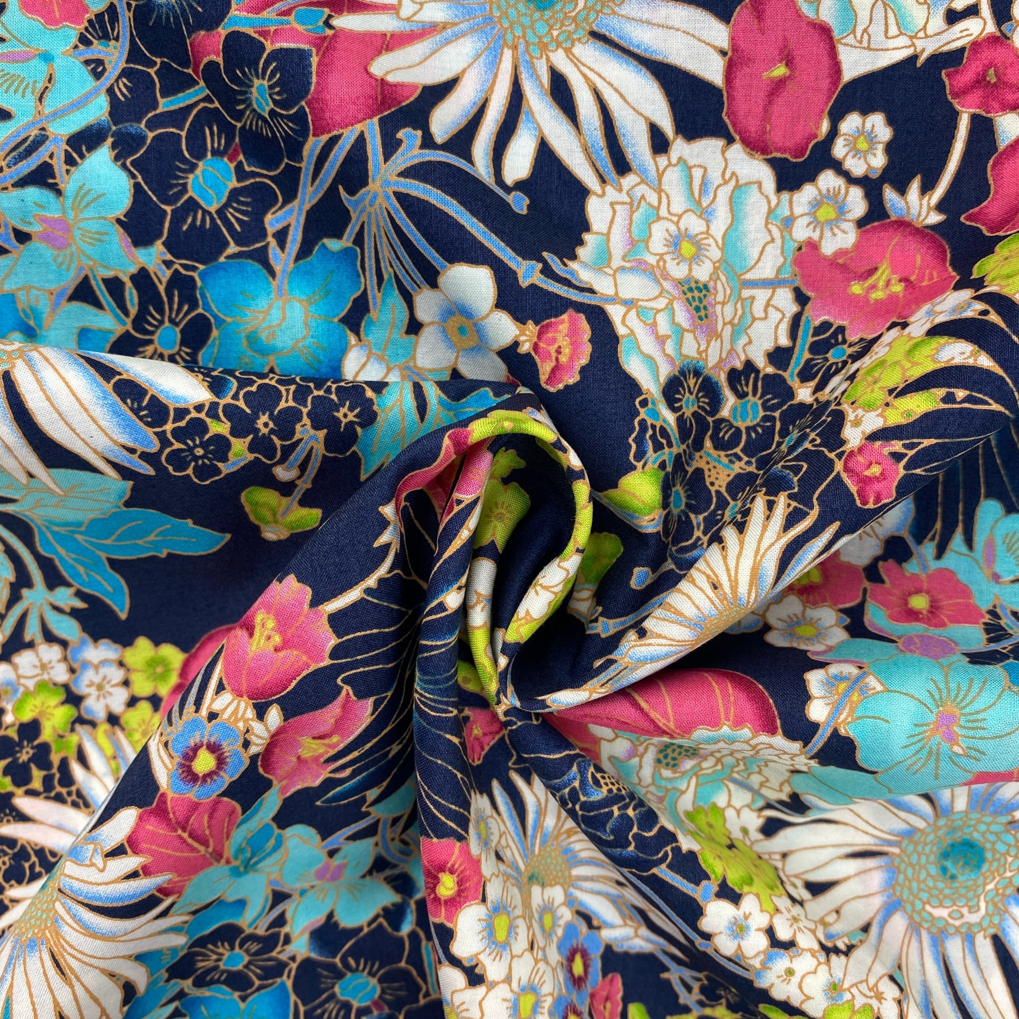 dressmaking fabric, cotton lawn, dark blue background, variety of flowers in barbie pink, apple green, sea blue, white, flowers outlined in a none shine gold. image is displayed flat with a scrunch in the middle.
