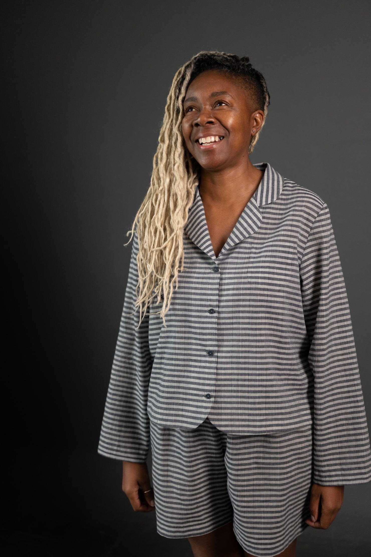 portrait image, merchant and mills Winnie pyjama pattern,woman standing, angled to right side, looking up, smiling. modelling long sleeve shirt, shorts, fabric is a light and dark grey stripe, background is black.