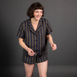 portrait image, merchant and mills Winnie pyjama pattern, woman stood, one foot unfront of the other, smiling, looking off camera, modelling short sleeve shirt, shorts, fabric is a navy and brown wide stripe, background is black.