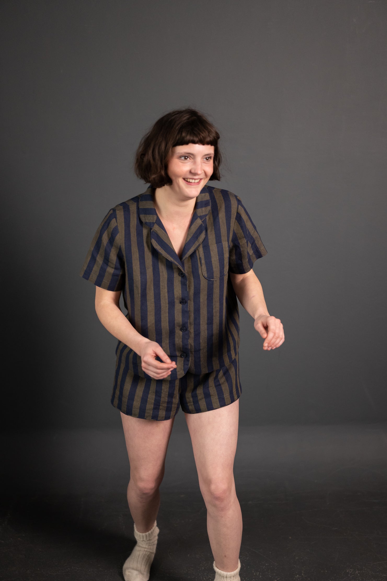 portrait image, merchant and mills Winnie pyjama pattern, woman stood, one foot unfront of the other, smiling, looking off camera, modelling short sleeve shirt, shorts, fabric is a navy and brown wide stripe, background is black.