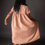 portrait image, merchant and mills Florence pattern, woman standing, facing back, looking ahead, right arm holding dressmodelling shin length dress, paired with white brogues, waist is gathered, bodice resembles a t-shirt styles. sleeves are short. fabric is peach background is black