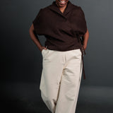 portrait image, black background, women stood face forward, smiling, hand in pockets of white cream wide fit trousers, top is a chocolate brown wrap with ties to the lefthand side.