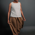 portrait image, black background, woman is stood angled eyes facing down, holding the side of her camel brown ankle length floaty skirts, paired with light grey linen vest top.
