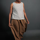 portrait image, black background, woman is stood angled eyes facing down, holding the side of her camel brown ankle length floaty skirts, paired with light grey linen vest top.