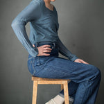 portrait image, grey studeio backdrop, woman sat on wooden stool, looking dow, hand on hip, modelling teal  long sleeve top, wearing deep blue jeans turned up at cuff.