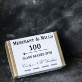 portrait image, black slate background, box angled to lean on wall, brown box, white sleeve, black text, merchant and mills 100 glass headed pins.