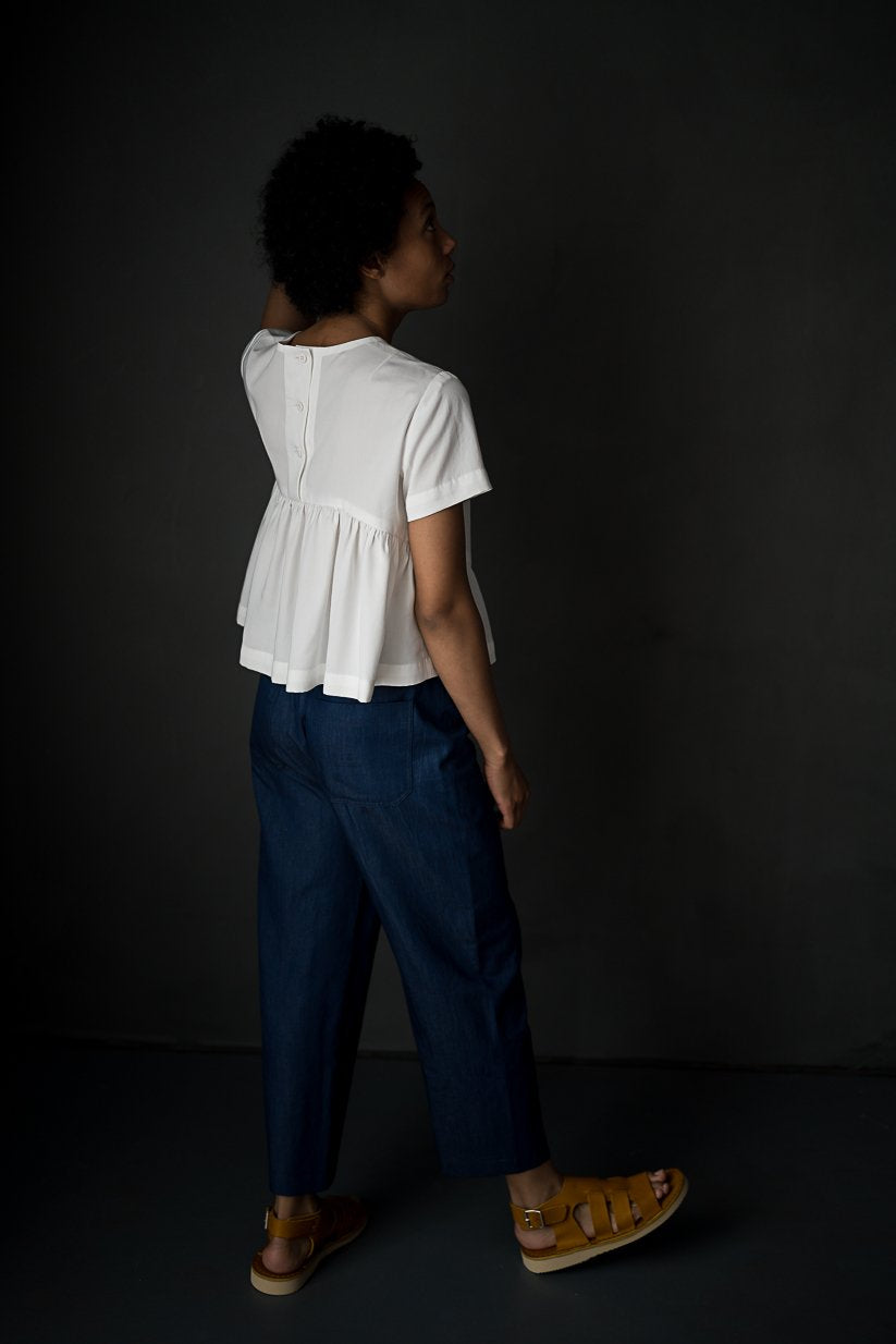portrait image, merchant and mills Florence pattern, woman standing, facing back, looking up, modelling hip length top, paired with denim blue trousers, waist is gathered, bodice resembles a t-shirt styles. sleeves are short. fabric is white background is black