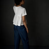 portrait image, merchant and mills Florence pattern, woman standing, facing back, looking up, modelling hip length top, paired with denim blue trousers, waist is gathered, bodice resembles a t-shirt styles. sleeves are short. fabric is white background is black