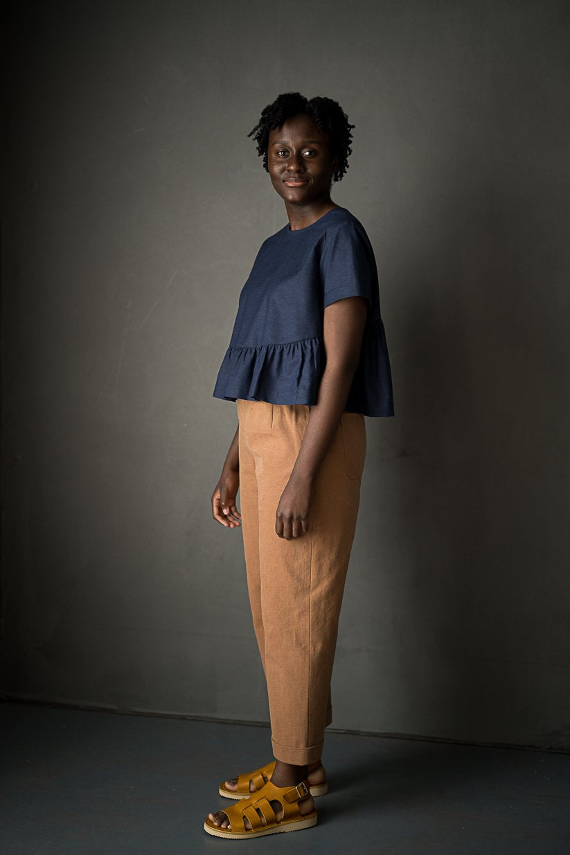 portrait image, black backdrop, woman stood tall, angled, looking at camera, modelling merchant and mill eve trousers, in a matt burnt orange fabric, paired with navy florence top.