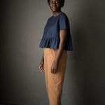 portrait image, black backdrop, woman stood tall, angled, looking at camera, modelling merchant and mill eve trousers, in a matt burnt orange fabric, paired with navy florence top.