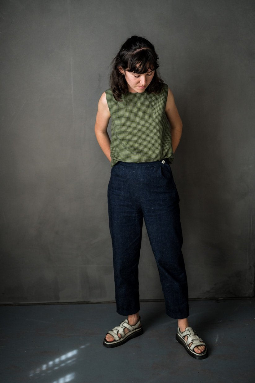 portrait image, black backdrop, woman stood smiling looking down, right hand slightky in pocket, left hand behind her back, modelling dark blue denim merchant and mill eve trouser pattern, paired with gingham green sleeveless top.