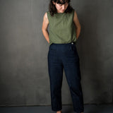 portrait image, black backdrop, woman stood smiling looking down, right hand slightky in pocket, left hand behind her back, modelling dark blue denim merchant and mill eve trouser pattern, paired with gingham green sleeveless top.