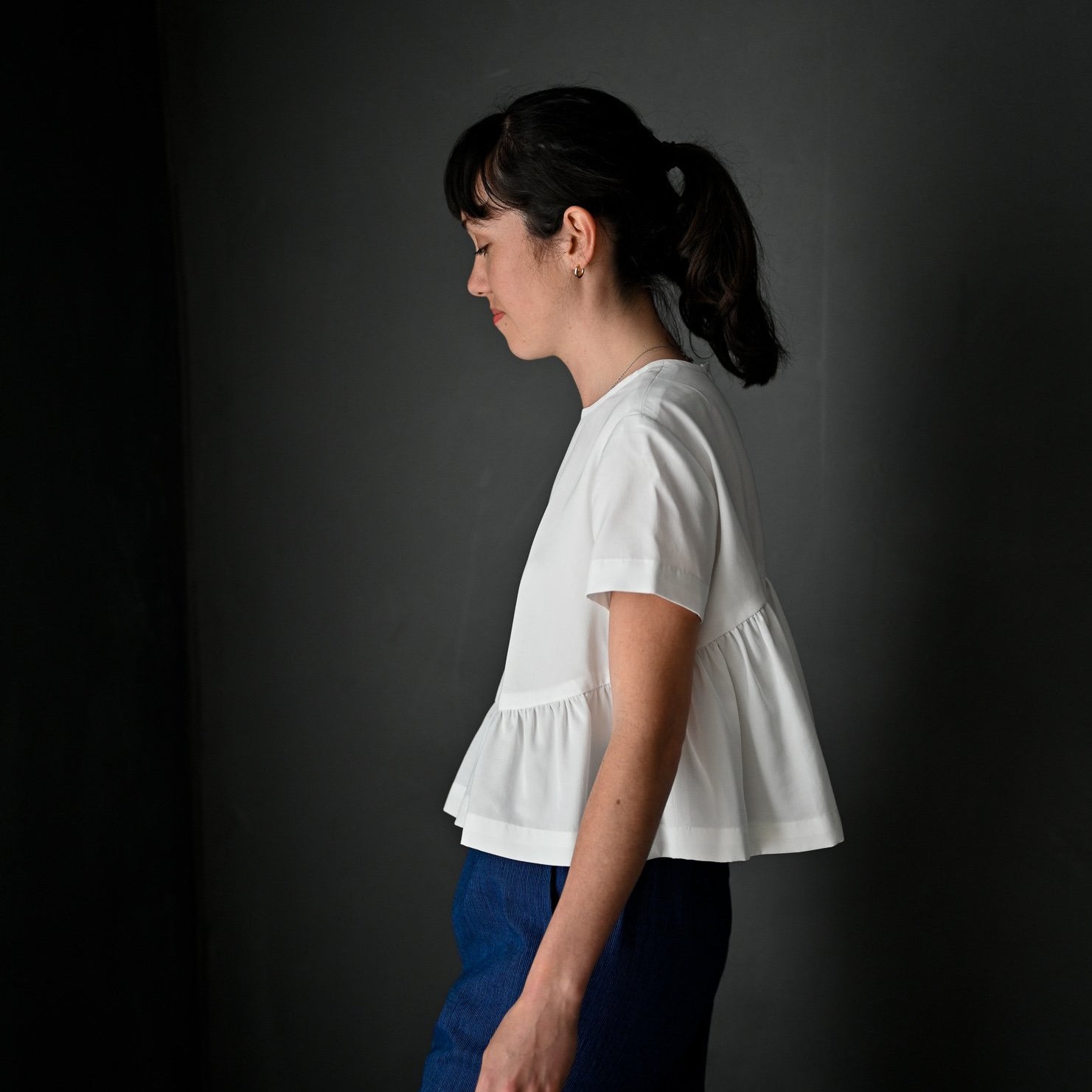 portrait image, merchant and mills Florence pattern, woman standing, facing side on, looking down, modelling hip length top, waist is gathered, bodice resembles a t-shirt styles. sleeves are short. fabric is white background is black