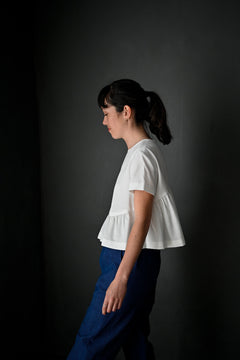 portrait image, merchant and mills Florence pattern, woman standing, facing side on, looking down, modelling hip length top, waist is gathered, bodice resembles a t-shirt styles. sleeves are short. fabric is white background is black